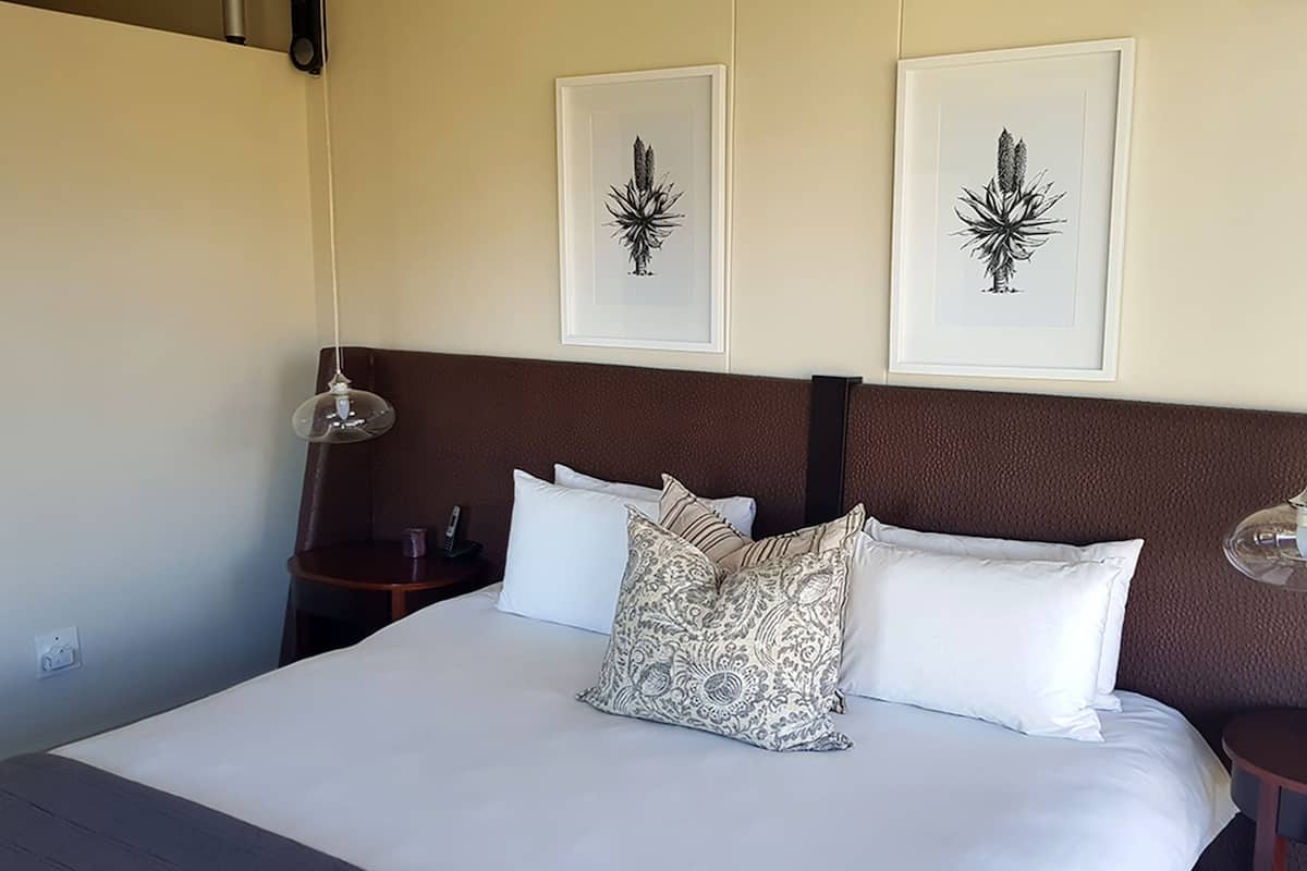 A neatly made bed with white linens and patterned pillows, a wooden headboard, and two framed botanical prints on the wall.