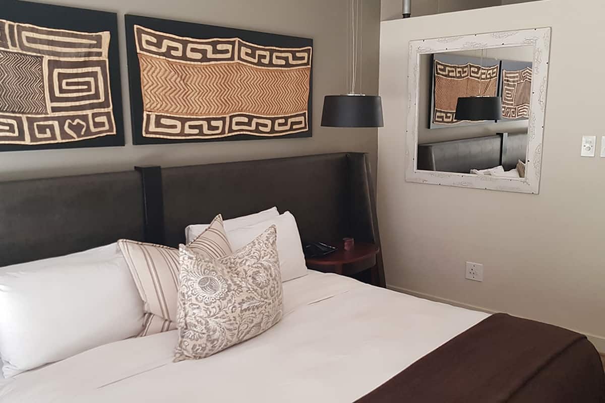 A neatly made bed with white linens and patterned pillows, a dark headboard, a bedside table with a lamp, and a mirror on the wall.