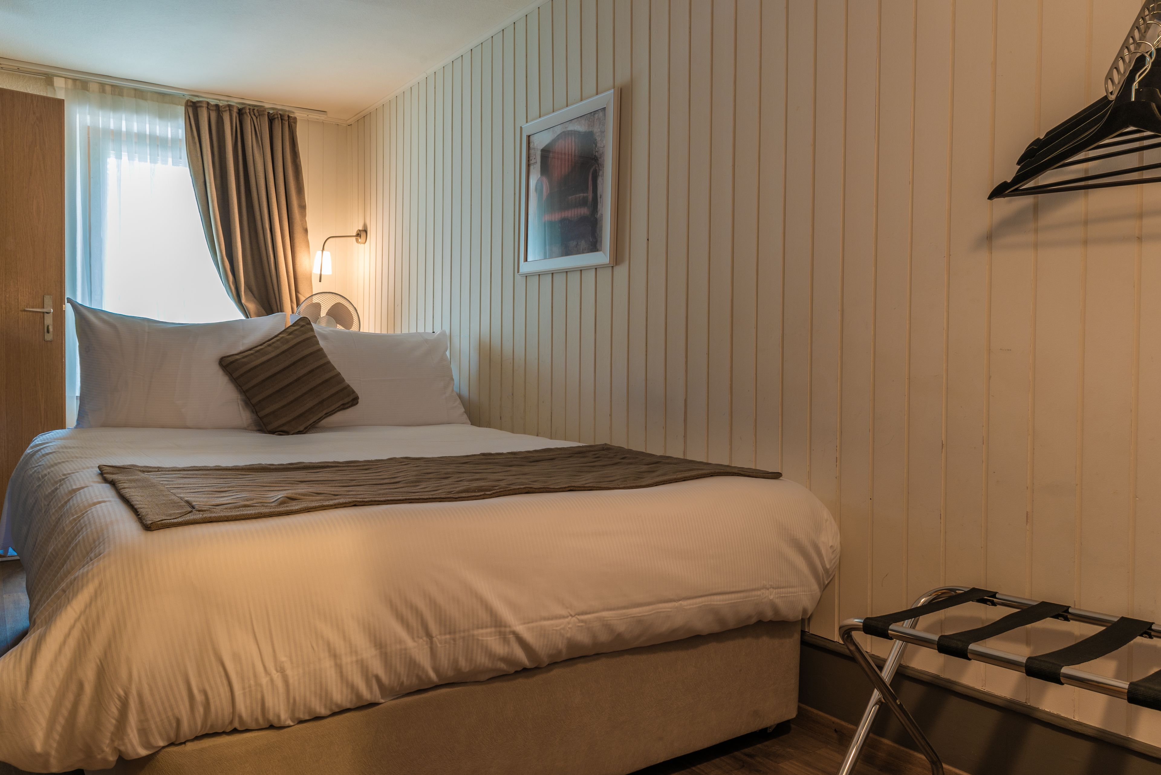 A neatly made bed with a pillow and a blanket, a bedside table with a lamp, and a window with curtains.
