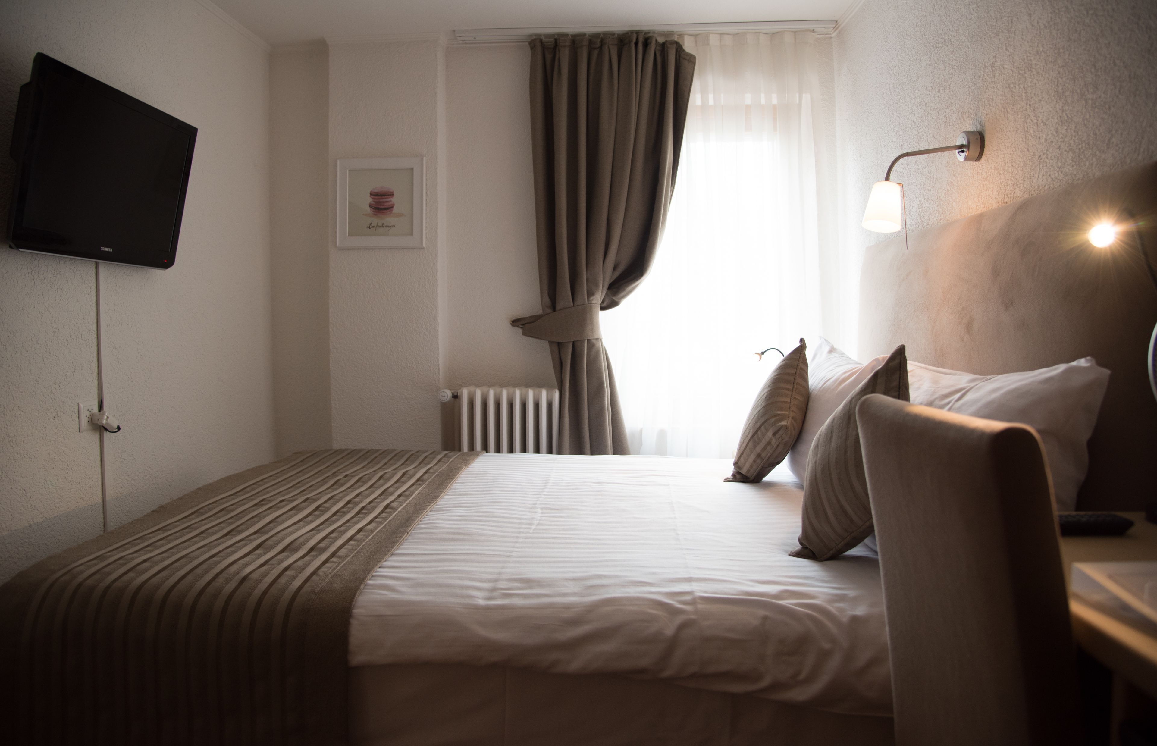 A hotel room with a bed, a TV mounted on the wall, a window with curtains, and a bedside lamp.