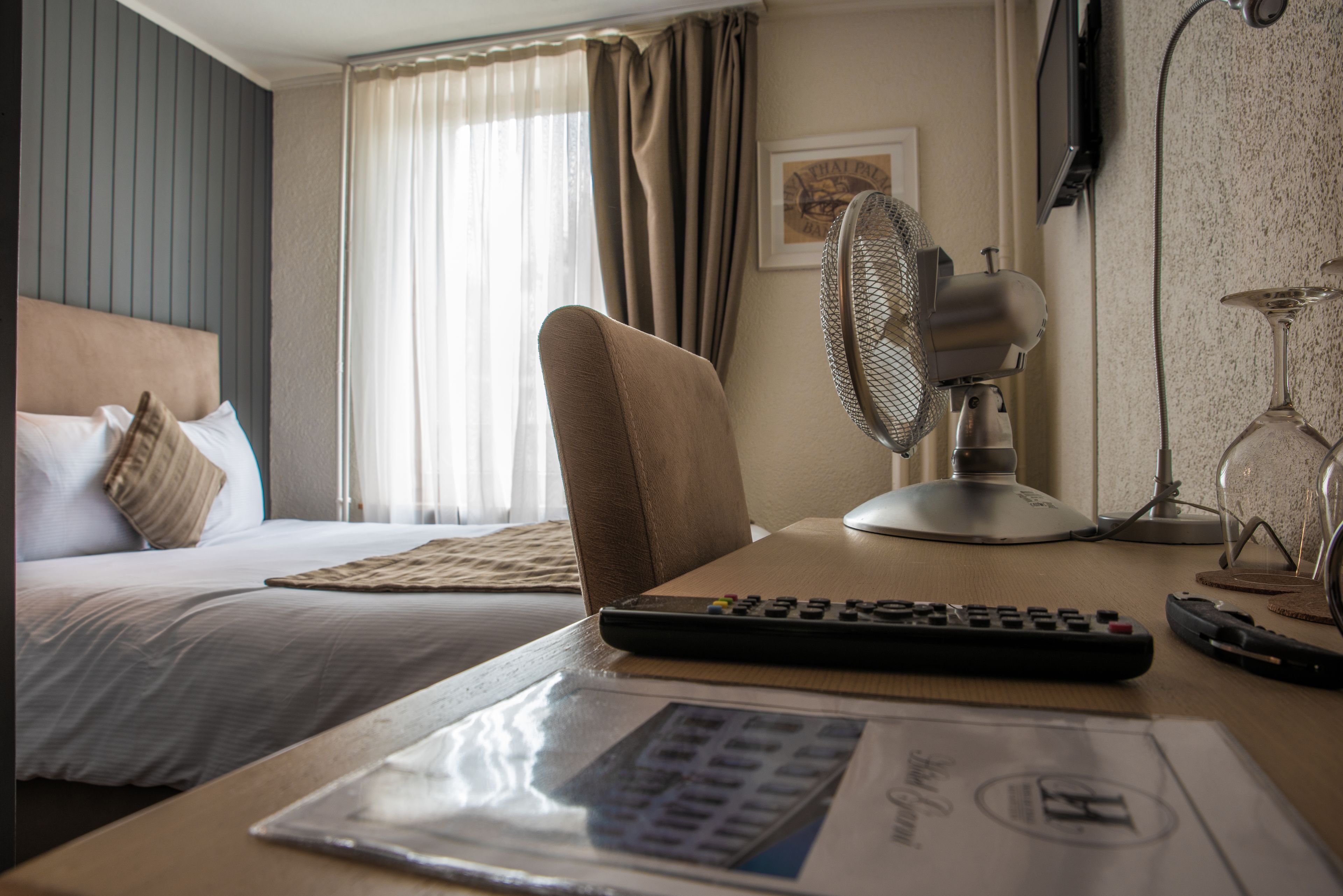 A hotel room with a bed, a desk with a remote control, a fan, and a lamp.