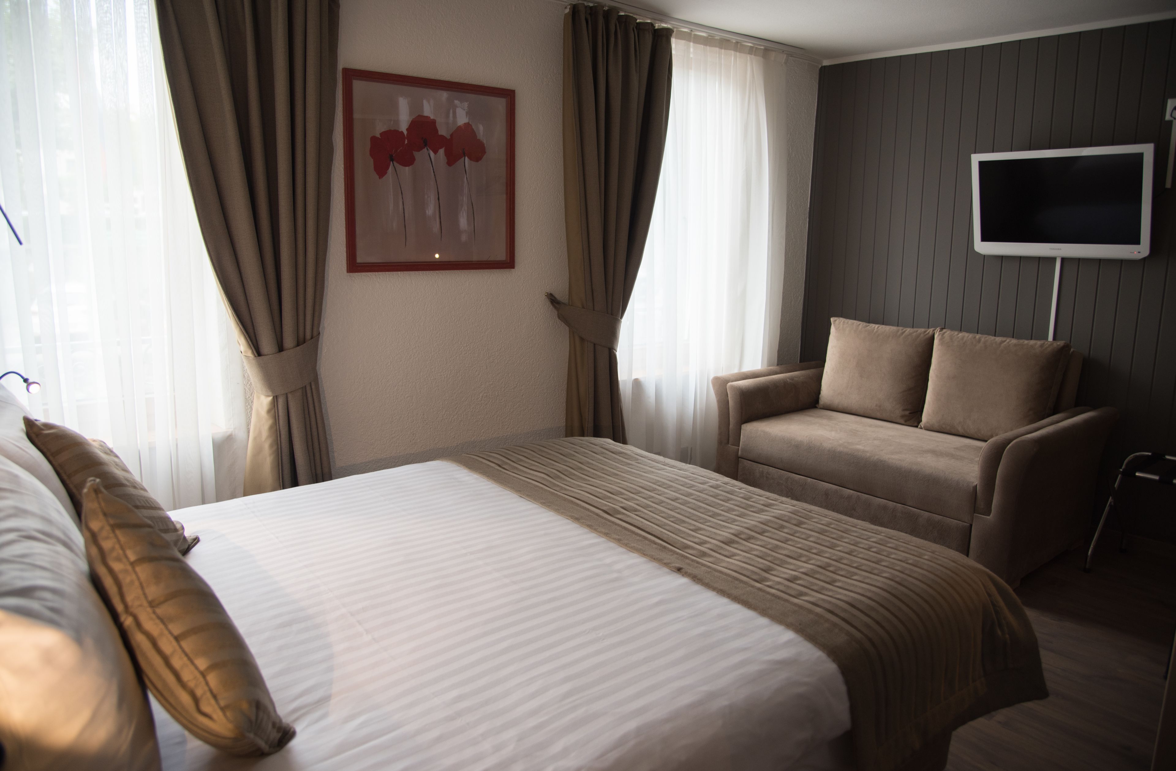 A hotel room with a large bed, a sofa, a TV mounted on the wall, and a framed artwork on the wall.