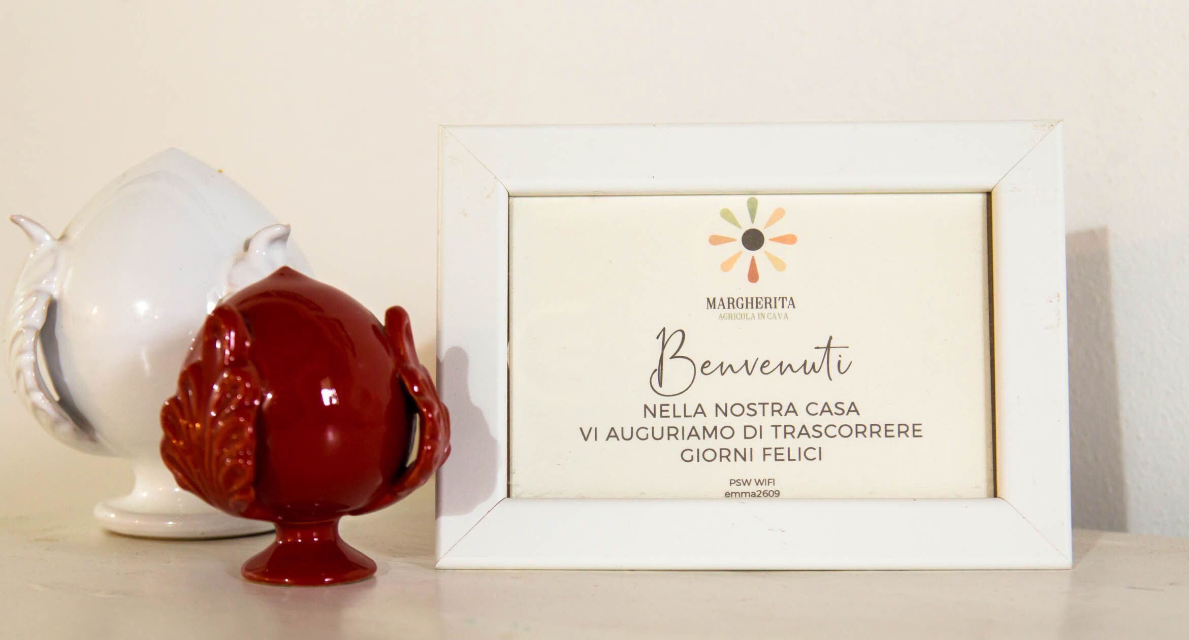Un cartell de benvinguda emmarcat amb text en italià, una tetera blanca i una tetera vermella.