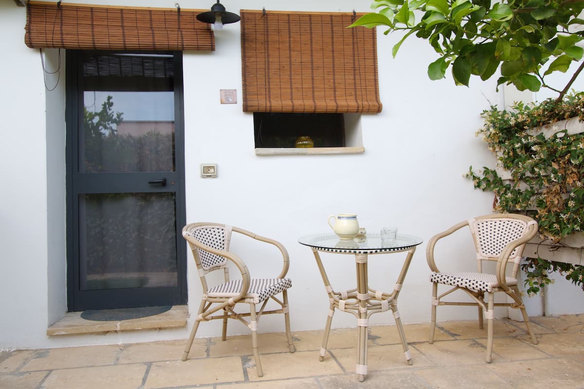 A small outdoor seating area with two wicker chairs and a glass table, a door with a black frame, and a window with a brown blind.