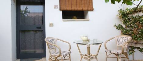A small outdoor seating area with two wicker chairs and a glass table, a door with a black frame, and a window with a brown blind.