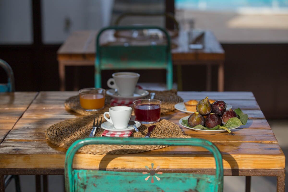 A wooden table with a woven placemat, a white cup, a glass of orange juice, a glass of red drink, and a plate of figs.