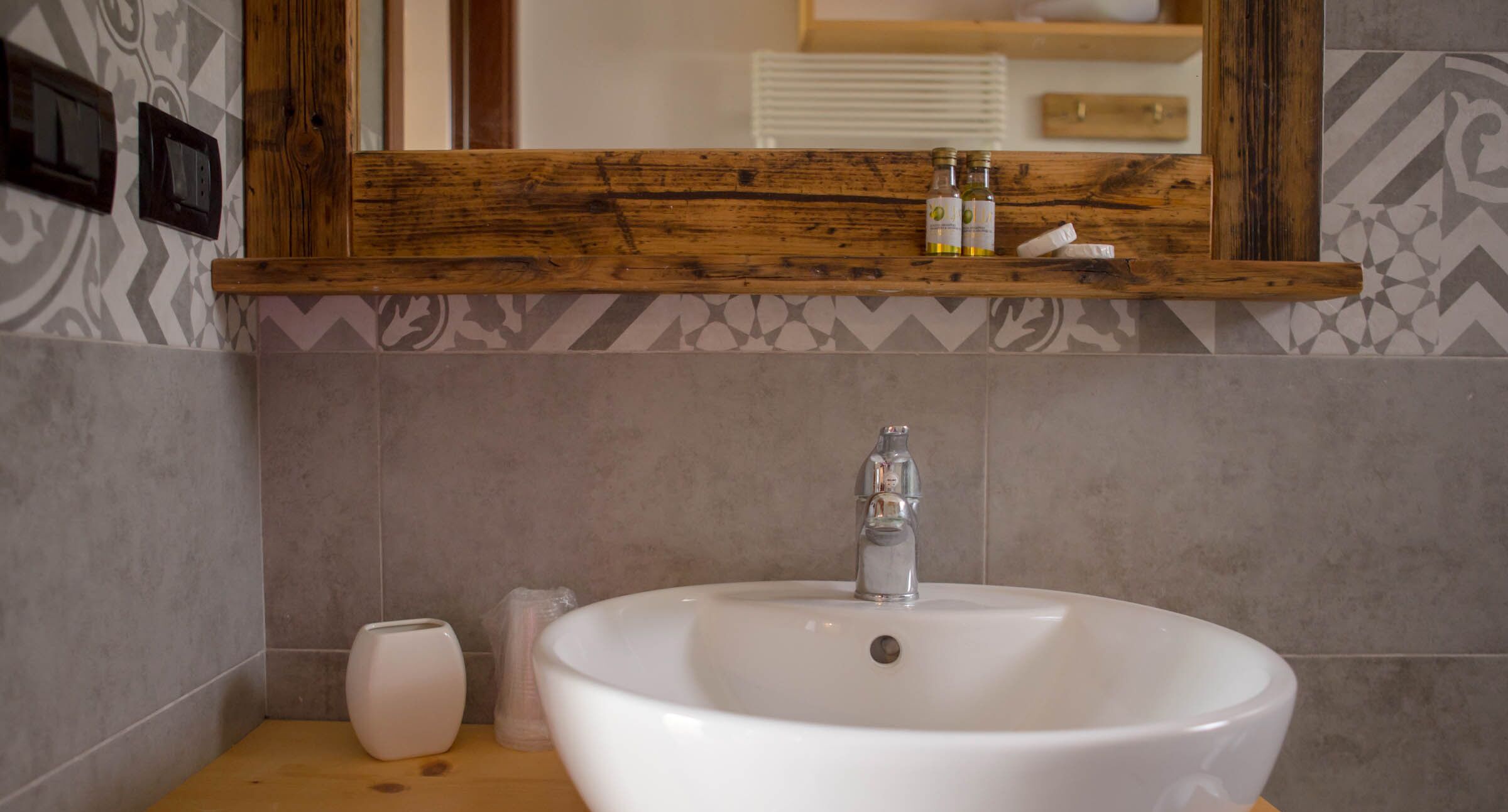 A bathroom with a white sink, a wooden shelf above it, and a mirror reflecting a bathroom setting.