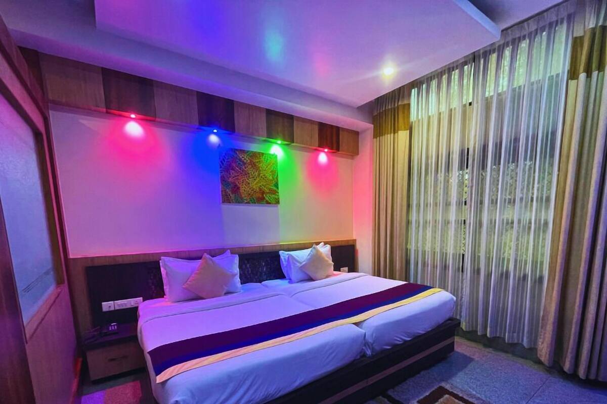 A modern hotel room with a large bed, a headboard with a colorful light strip, a large window with curtains, and a small bedside table.