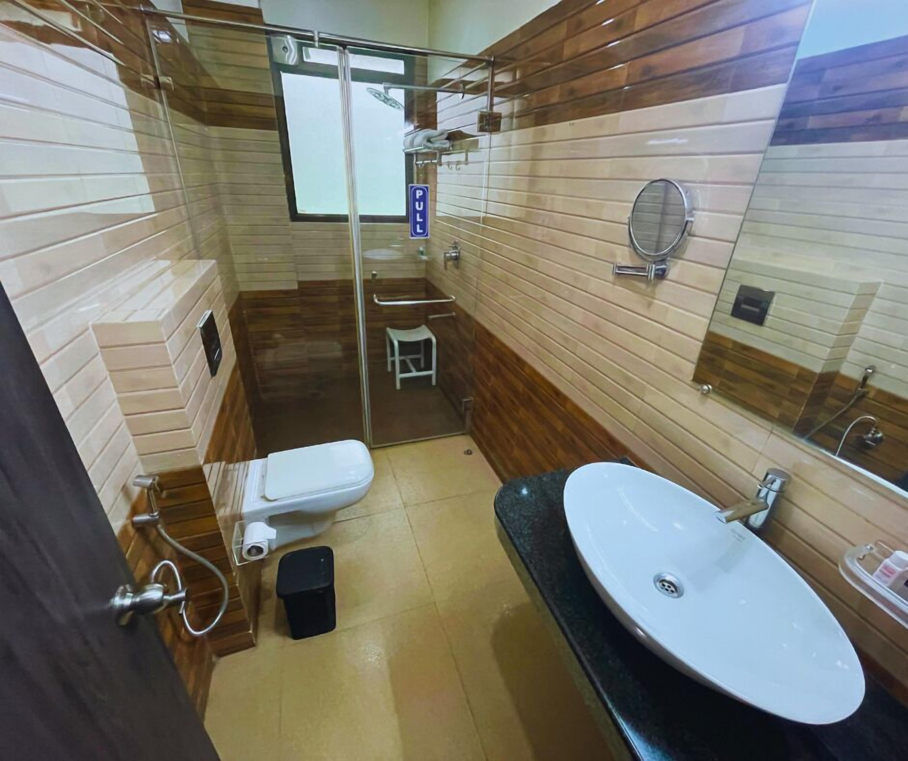 A bathroom with a glass-enclosed shower, a white toilet, a sink with a mirror, and a small stool.