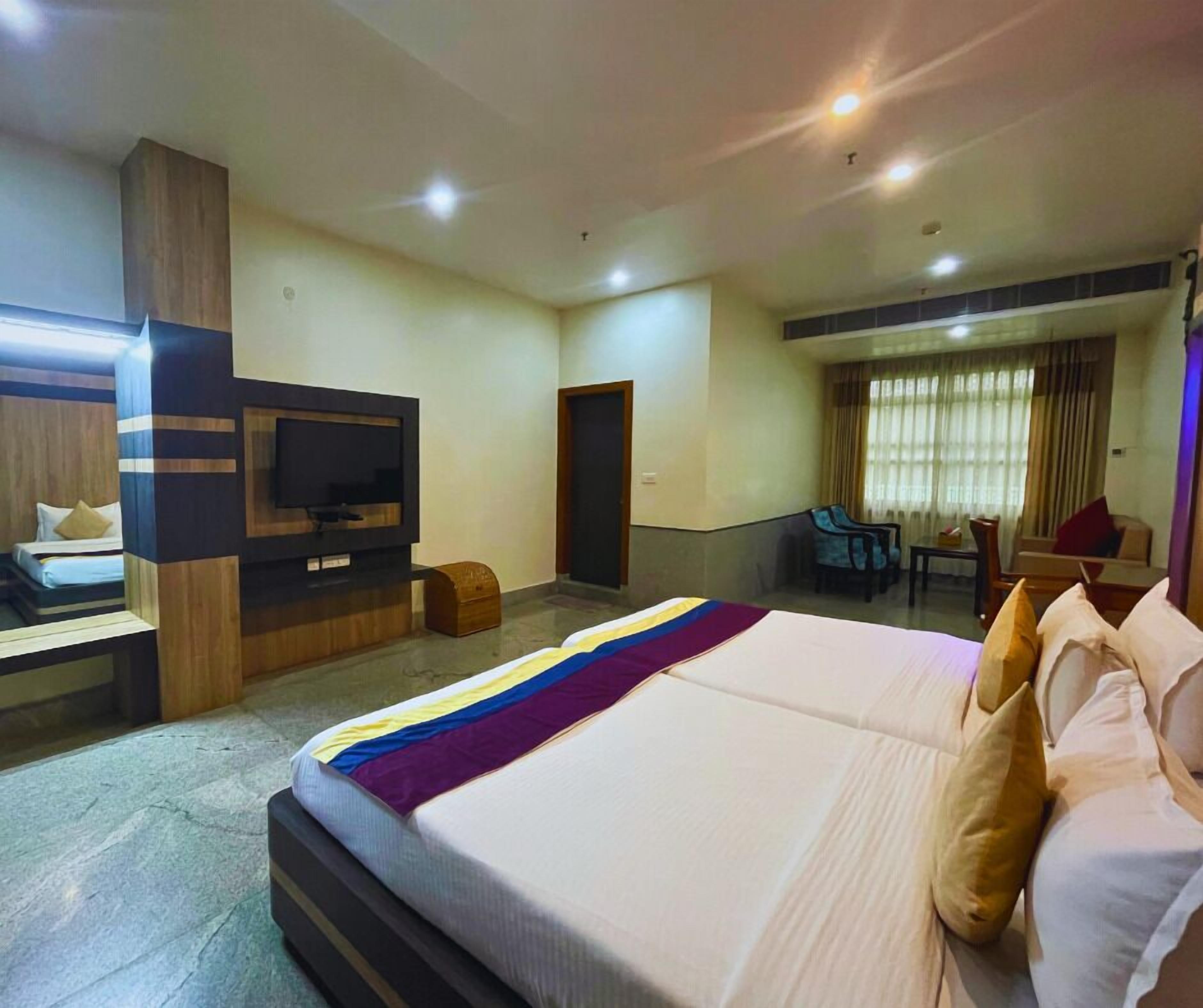 A hotel room with a large bed, a TV mounted on the wall, a desk with a chair, and a window with curtains.