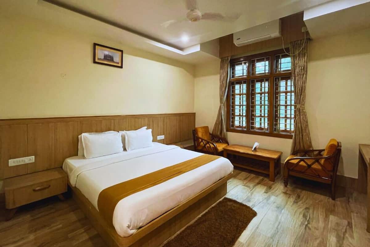 A hotel room with a large bed, a desk, two chairs, a window with curtains, an air conditioning unit, and a ceiling fan.
