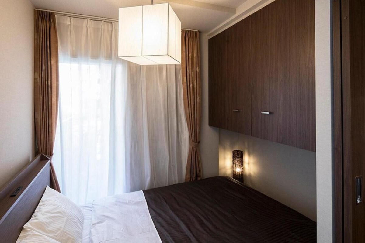 A hotel room with a bed, a bedside table, a nightstand, a wall-mounted lamp, and a wardrobe.