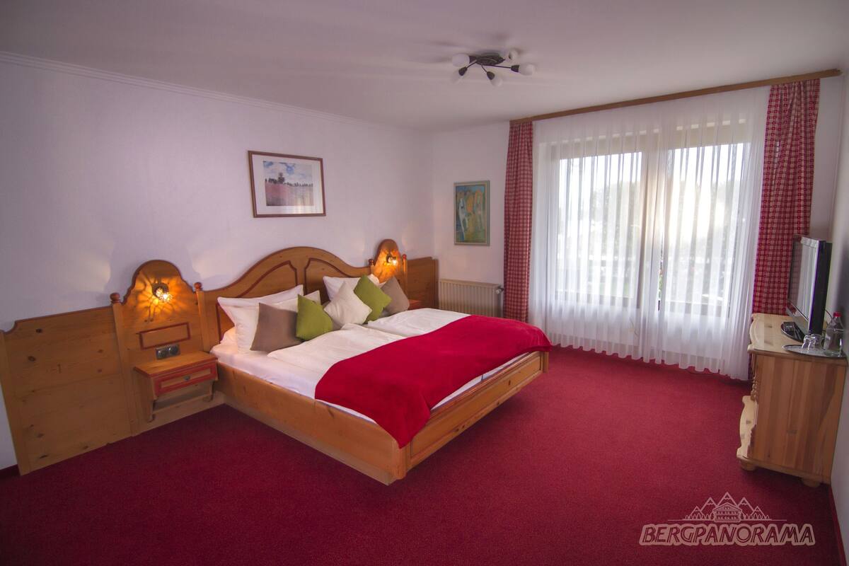 A bedroom with a wooden bed, red carpet, a nightstand, a bedside table, a ceiling fan, and a window with curtains.
