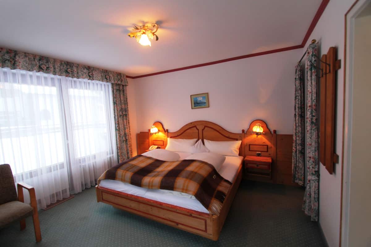 A bedroom with a wooden bed, two bedside tables with lamps, a chair, and a window with sheer curtains.