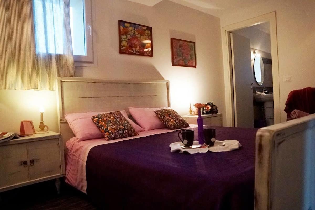 A bedroom with a bed, bedside table, and a nightstand. There is a window with curtains, a door, and a mirror.