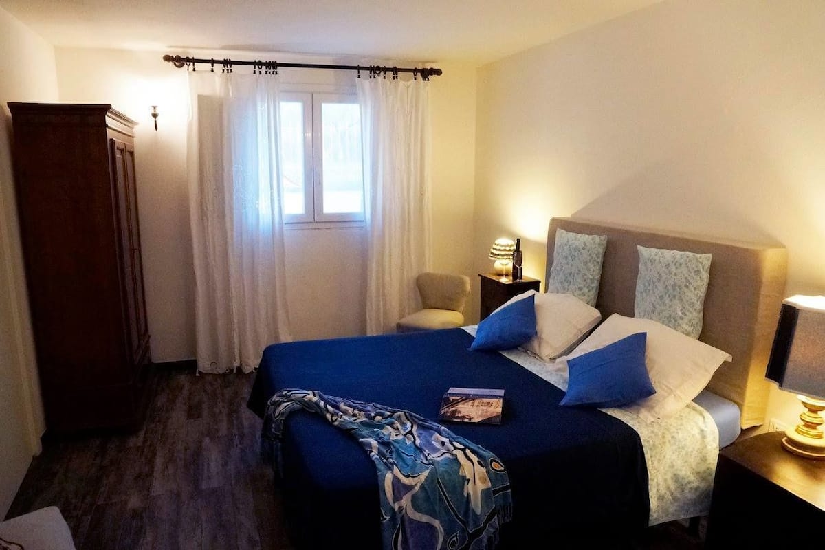 A hotel room with a large bed, a wardrobe, a window with curtains, a bedside table with a lamp, and a nightstand.