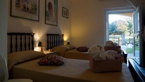 A hotel room with two beds, a bedside table with lamps, a window with a view of the outside, and a basket of towels. - Molino Della Volta (Grosseto)