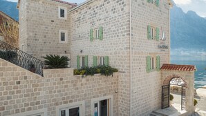 A stone building with green shutters, a balcony with plants, and a view of the sea.