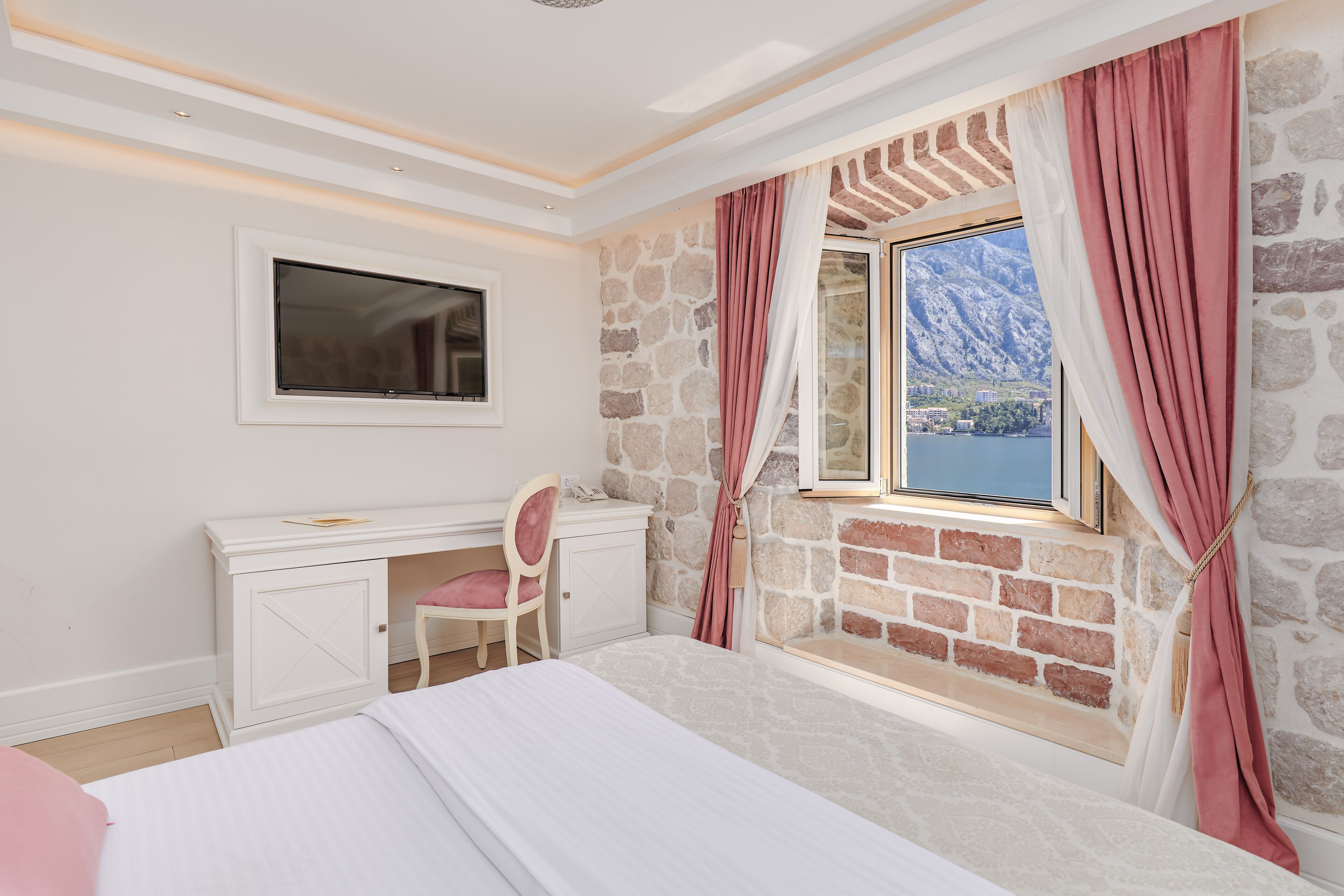 Um quarto com cama, escrivaninha, cadeira, janela com cortinas e parede de pedra.