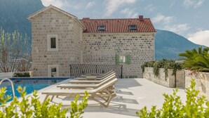 A stone building with a tiled roof, a pool, and lounge chairs.