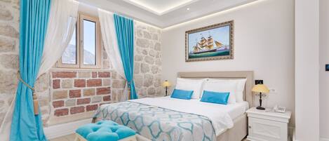A bedroom with a stone wall, a bed with blue and white bedding, a nightstand with a lamp, and a painting of a ship on the wall.