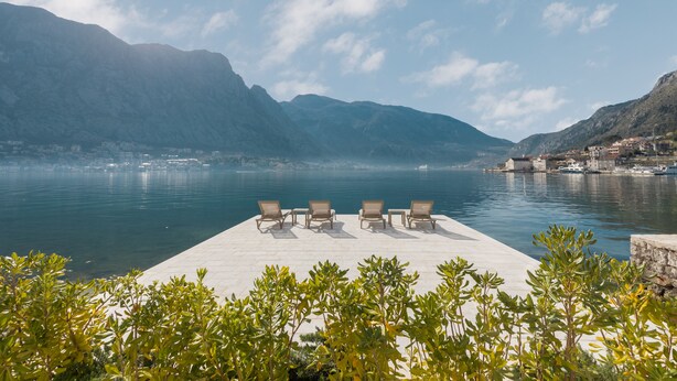 A waterfront terrace with lounge chairs, a clear view of the lake, and mountains in the background.