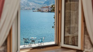 A view from a window overlooking a lake with a building on the opposite shore, two chairs, and a small table.