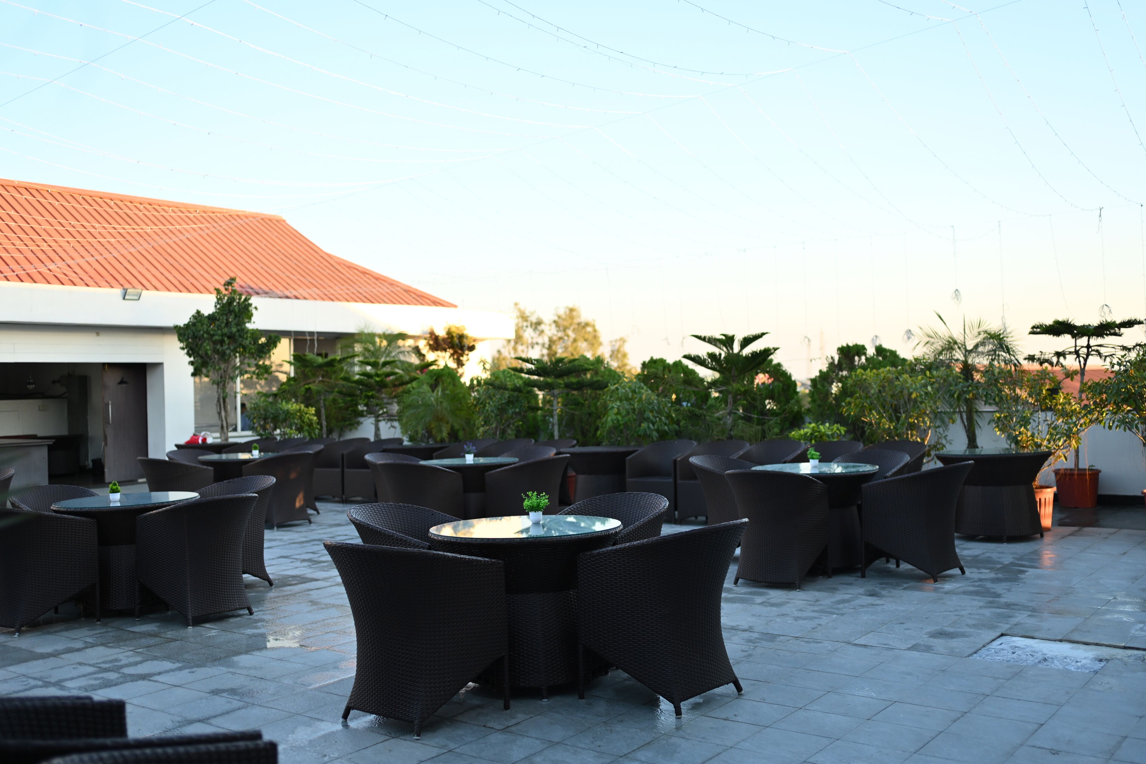 An outdoor patio area with multiple round tables and wicker chairs, surrounded by potted plants and a building with a red roof in the background.