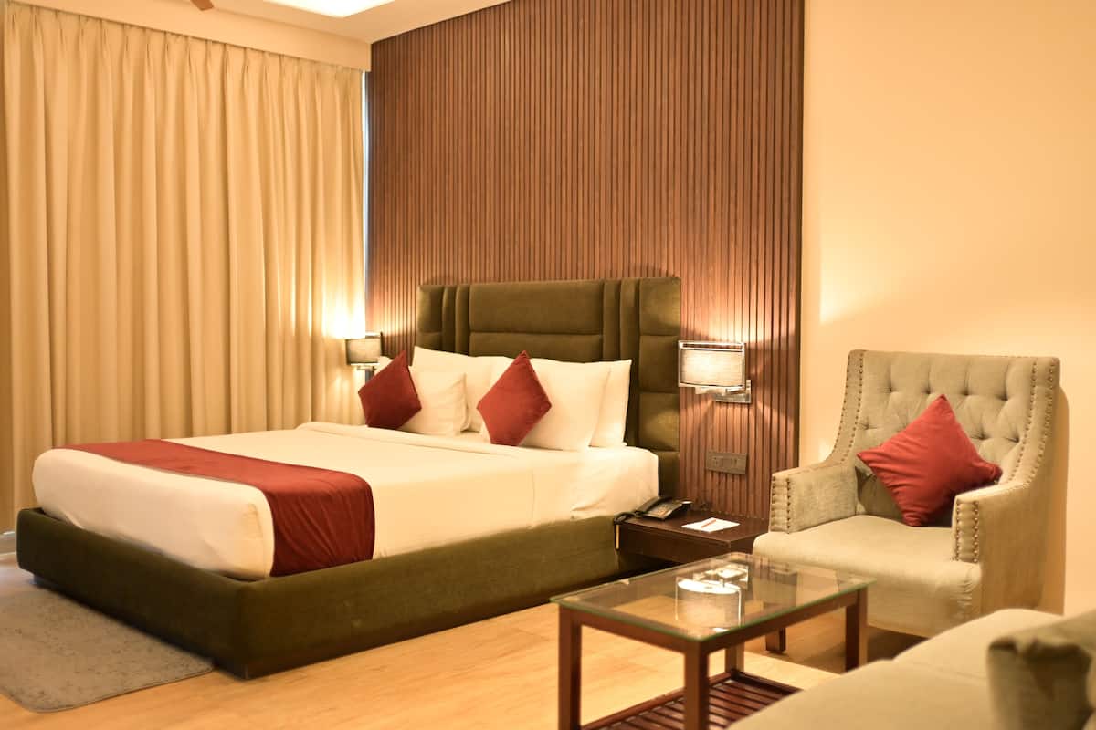 Habitación de hotel con una cama grande, cabecera tapizada de verde, una mesa de centro de vidrio, una mesita de noche de madera y un cojín decorativo rojo.