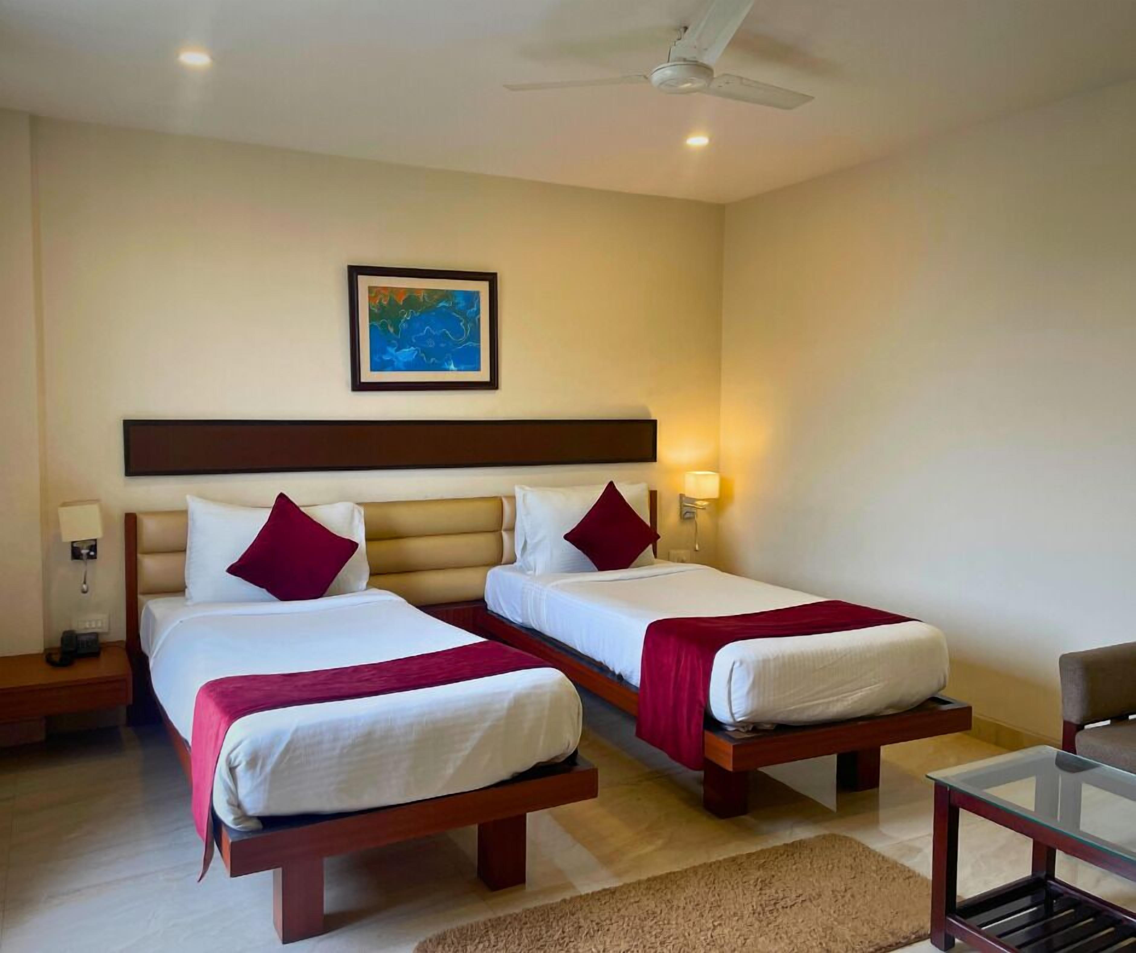 A hotel room with two single beds, a ceiling fan, a nightstand with a lamp, a framed picture on the wall, and a small table with a chair.