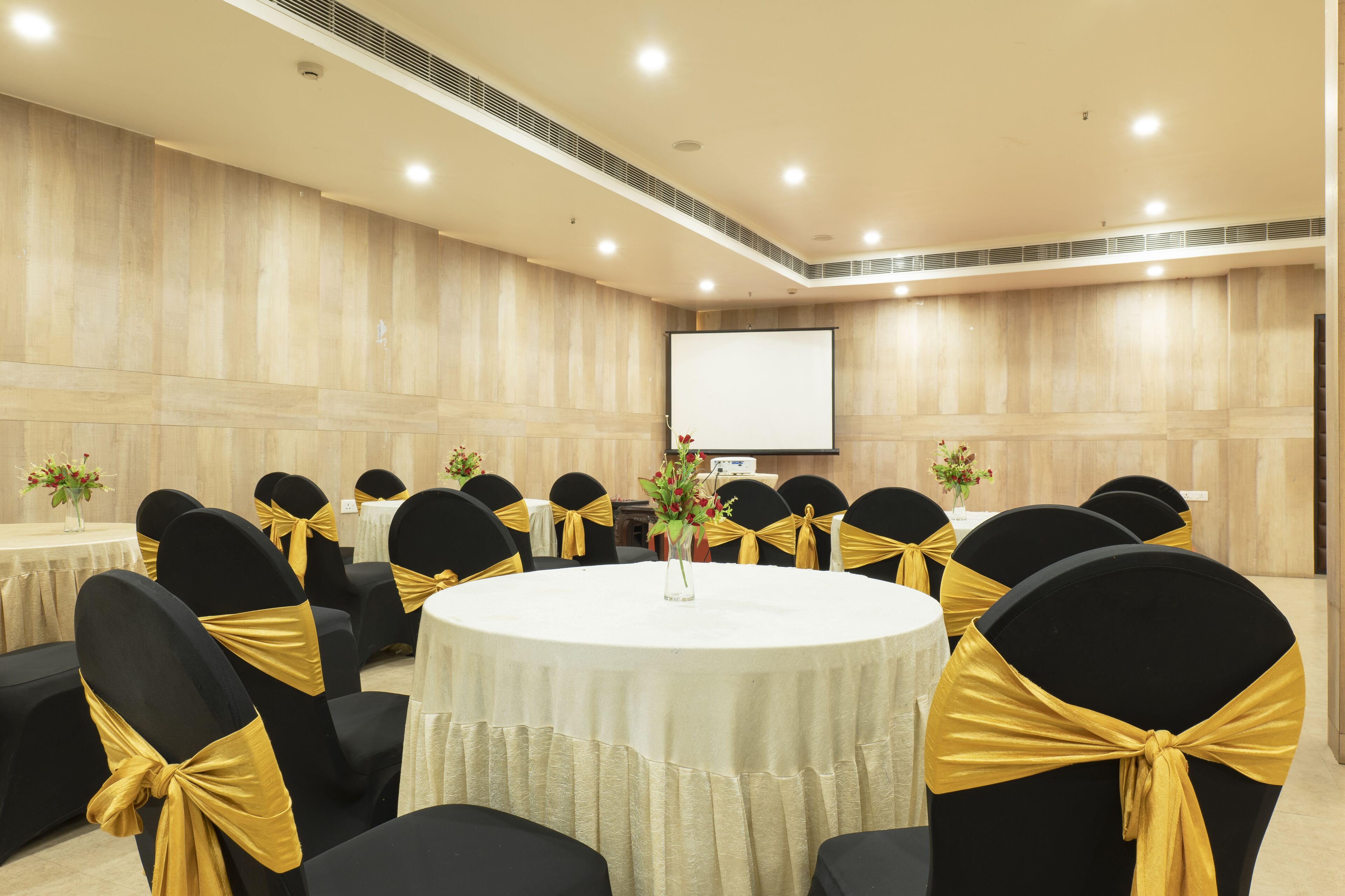 Uma sala de conferências com mesas redondas, cadeiras pretas com fitas douradas e uma tela de projetor na parede.