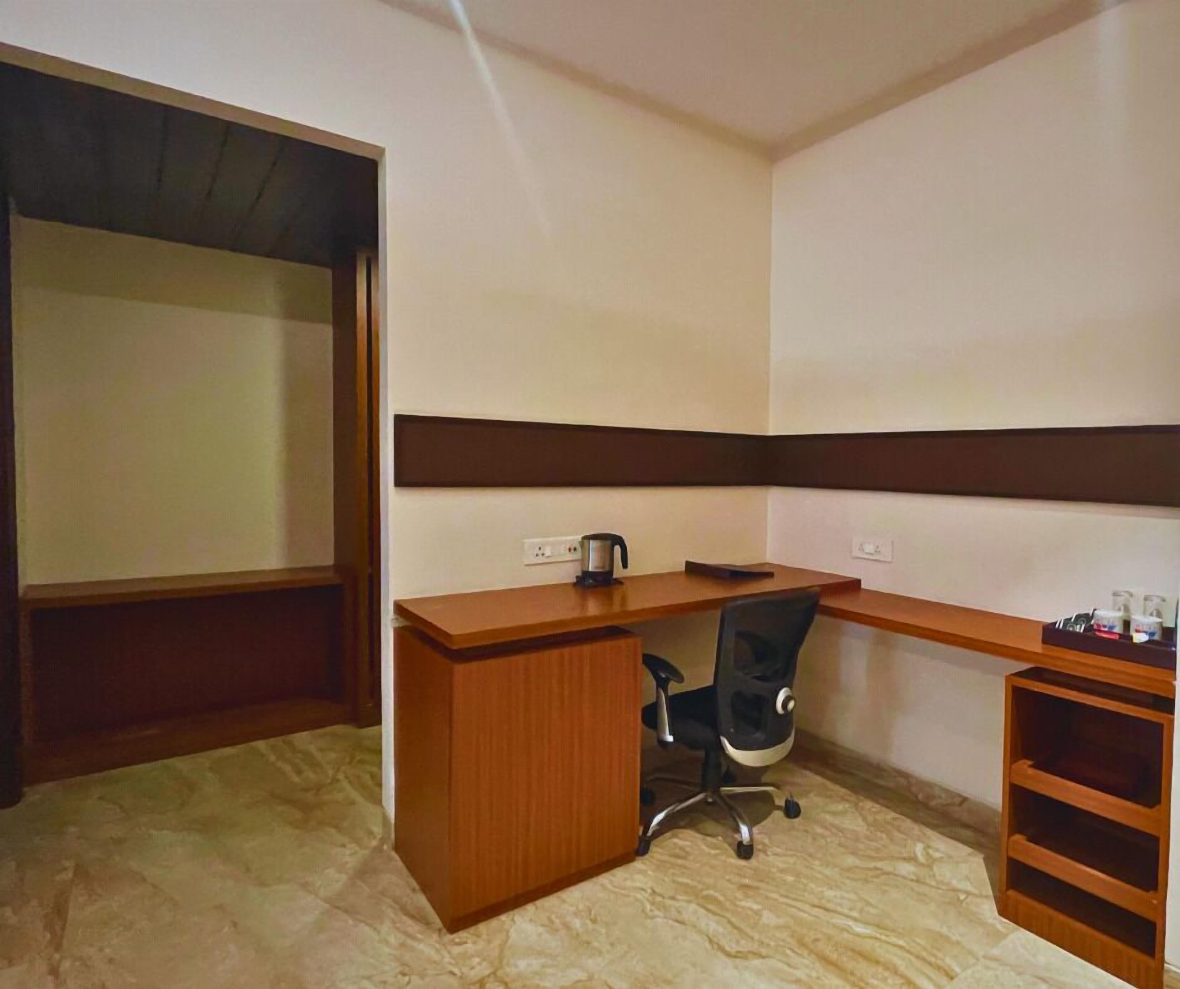 A small office space with a wooden desk, a black office chair, a kettle, and a shelf with bottles.