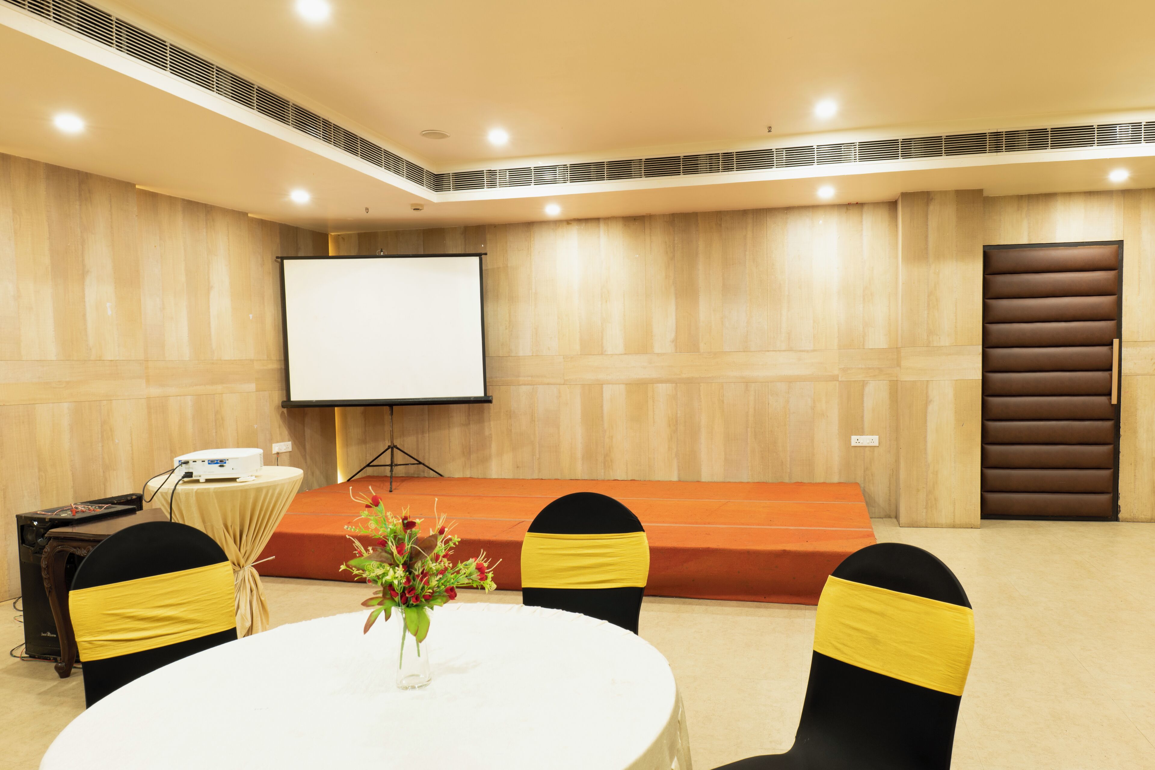 Un salón de conferencias con una mesa blanca, sillas negras con cintas amarillas, una pantalla de proyector y un atril.