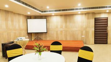 Un salón de conferencias con una mesa blanca, sillas negras con cintas amarillas, una pantalla de proyector y un atril.