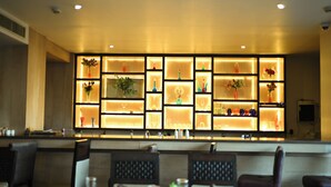 Zona de bar con estanterías iluminadas que exhiben diversos vasos y botellas. Hay arreglos de asientos con mesas y sillas frente a la barra.