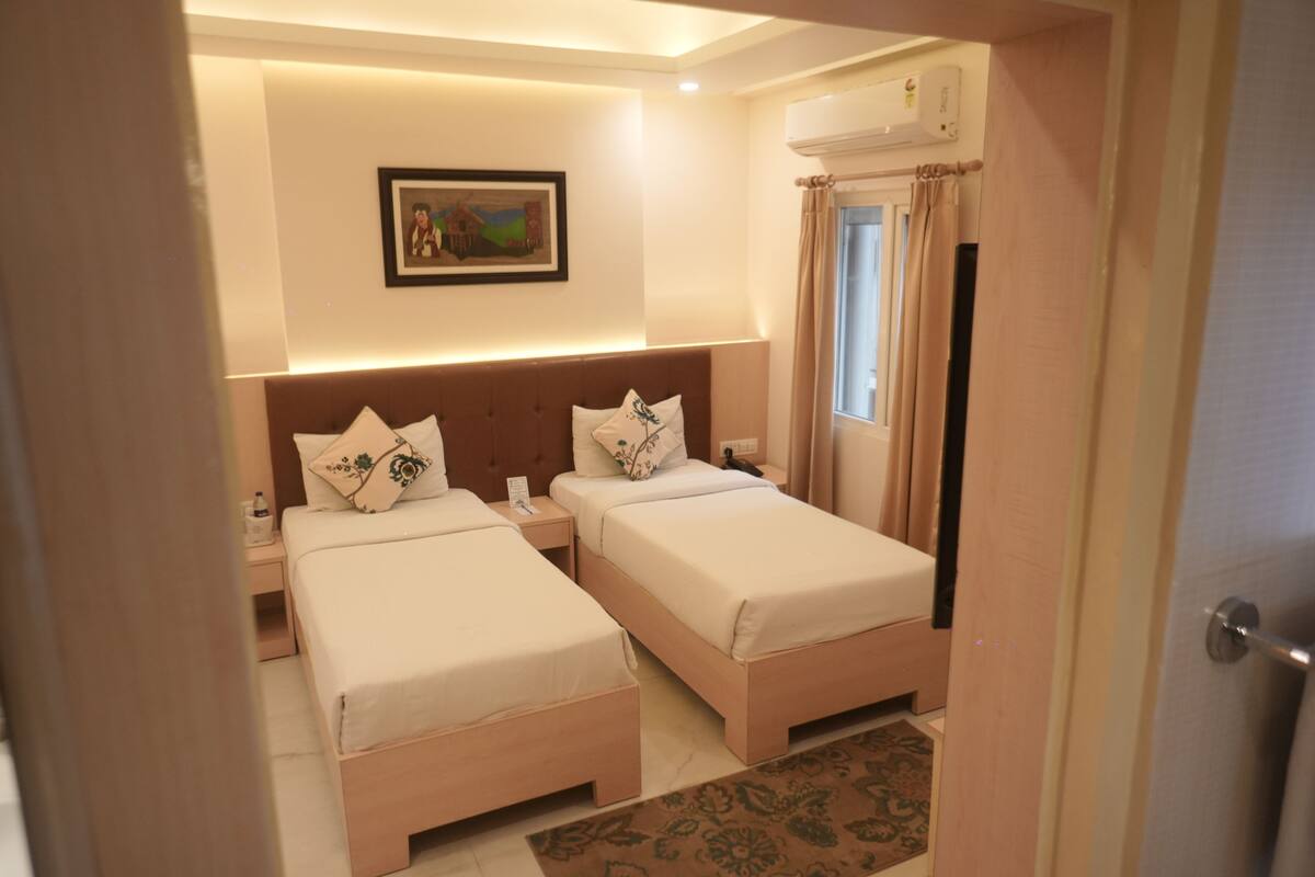 A hotel room with two beds, a nightstand, a wall-mounted air conditioner, a framed picture, and a window with curtains.
