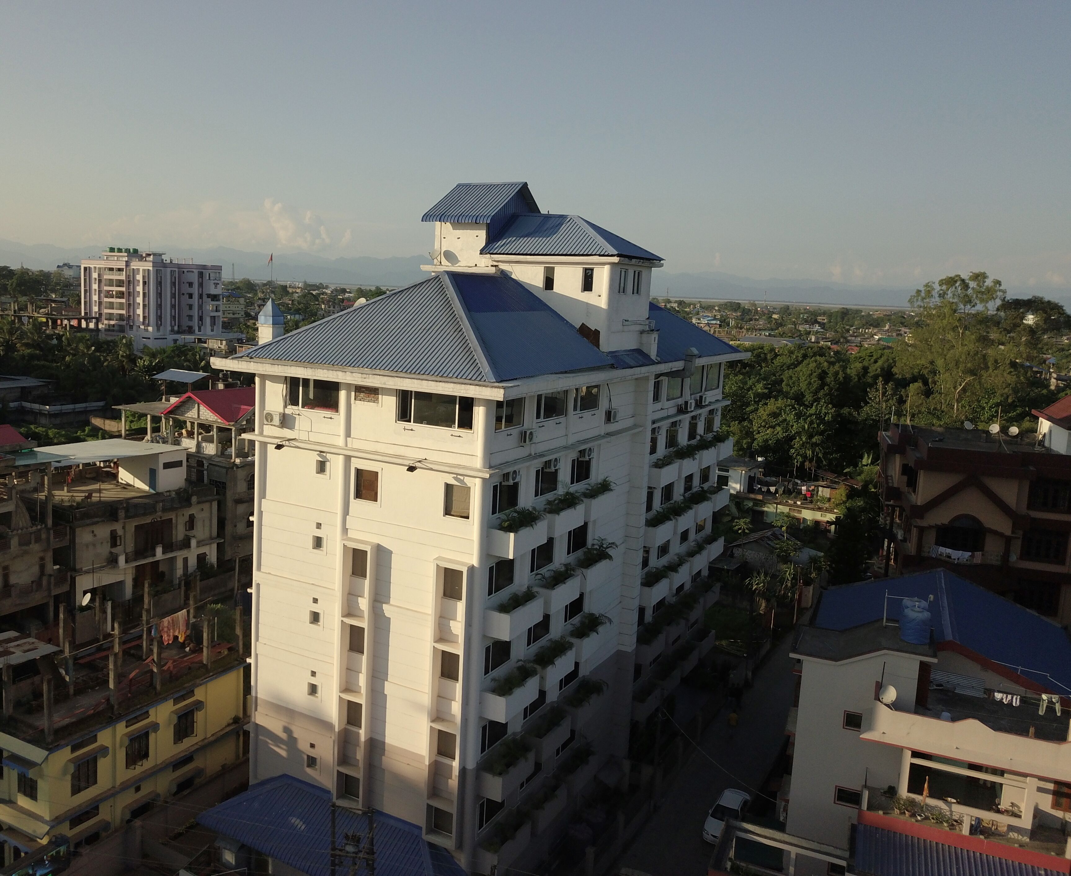 Un edificio de varios pisos con techo plano y balcones, rodeado de otros edificios y vegetación.