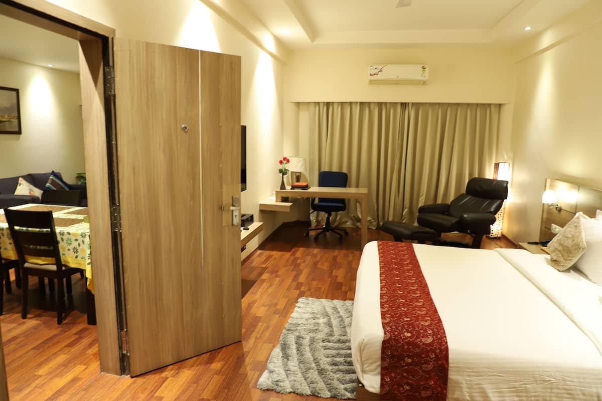 A hotel room with a wooden wardrobe, a desk with a chair, a dining area with a table and chairs, and a bed with a red and white bedspread.
