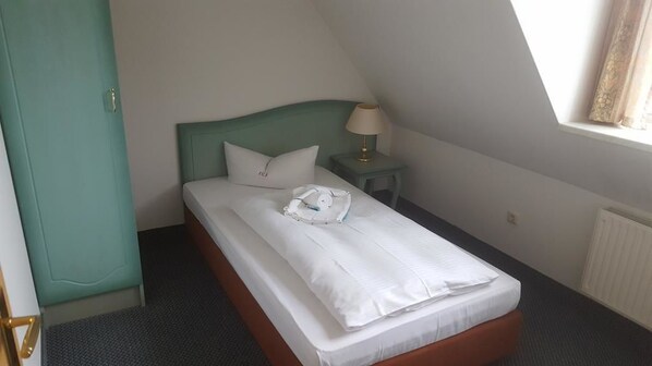 Una cama individual con ropa de cama blanca, cabecera verde, mesita de noche con lámpara y ventana con cortinas.