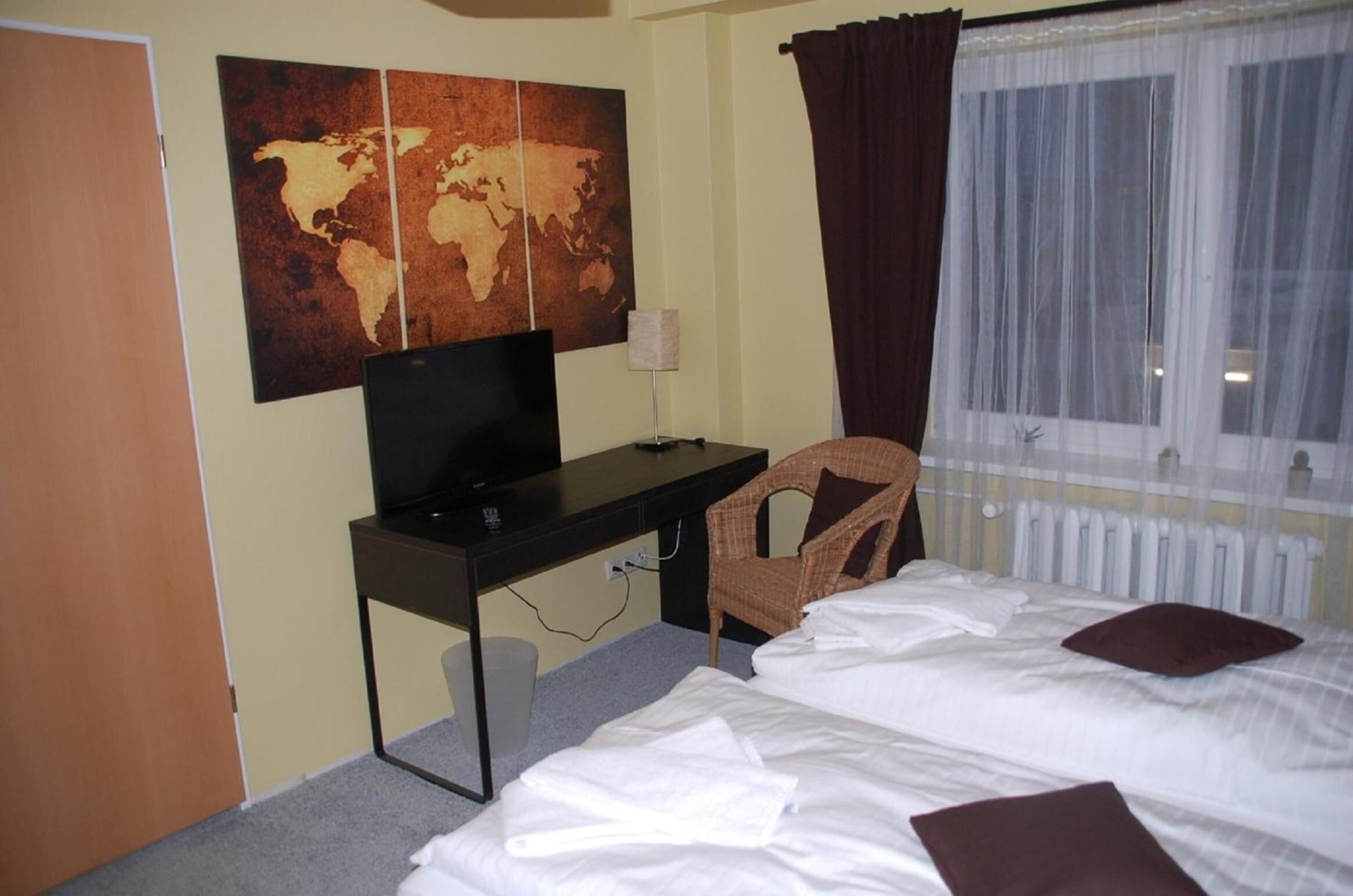 Quarto de hotel com cama, escrivaninha com computador, cadeira, mapa-múndi na parede e janela com cortinas.