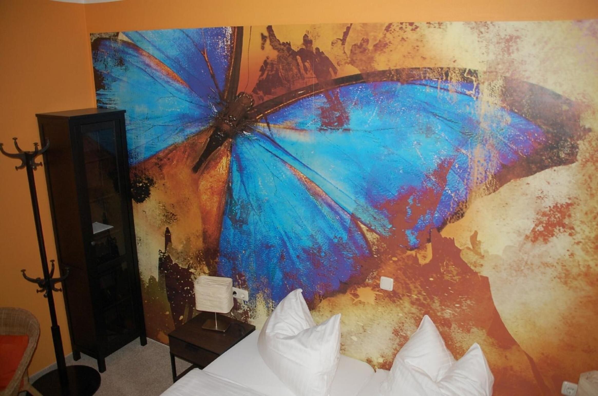 A room with a large abstract painting of a blue butterfly on the wall, a black cabinet, a small table with a white pillow, and a chair.