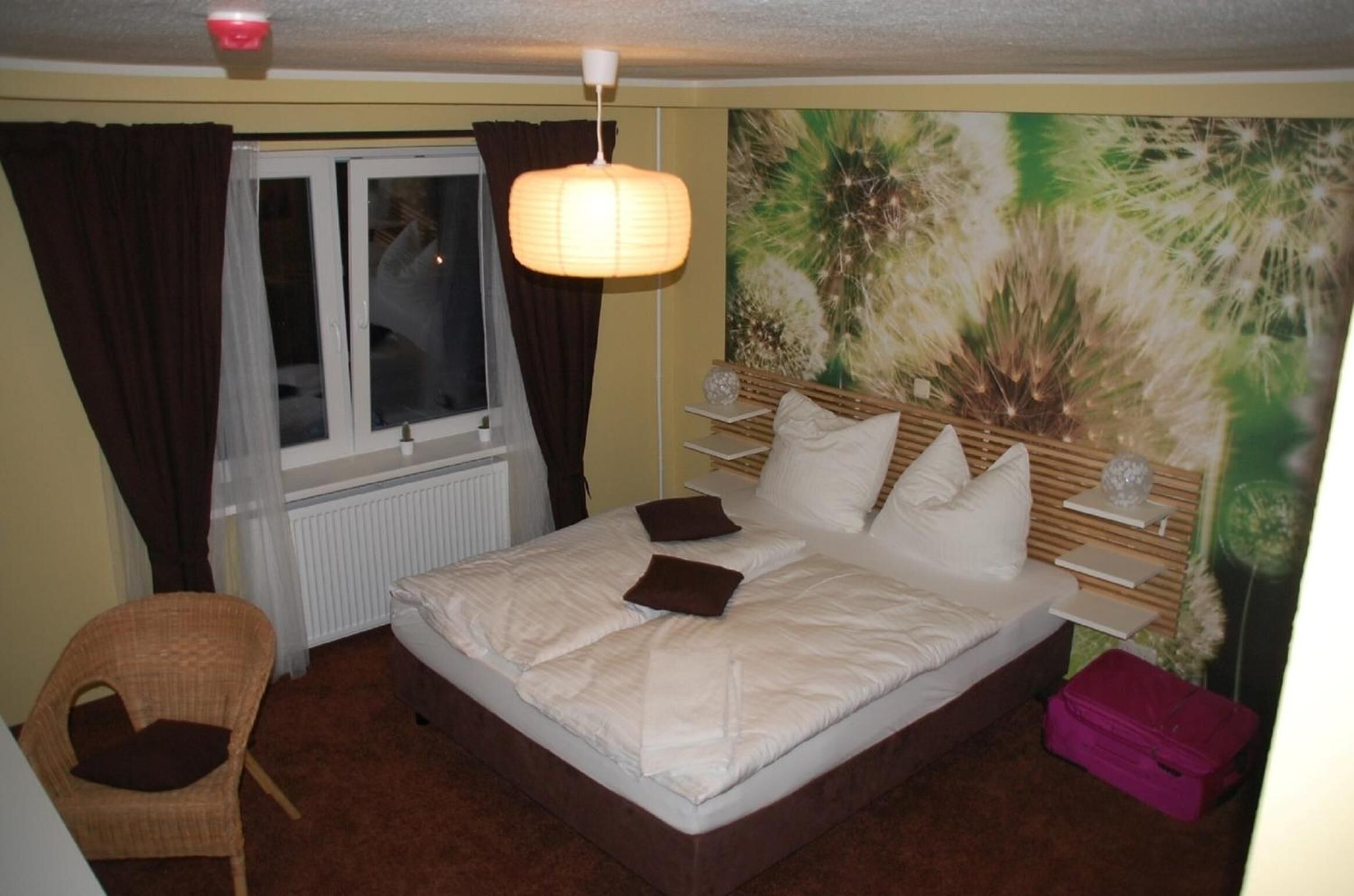 A hotel room with a bed, a chair, a window, a radiator, a wall painting, and a nightstand with a lamp.