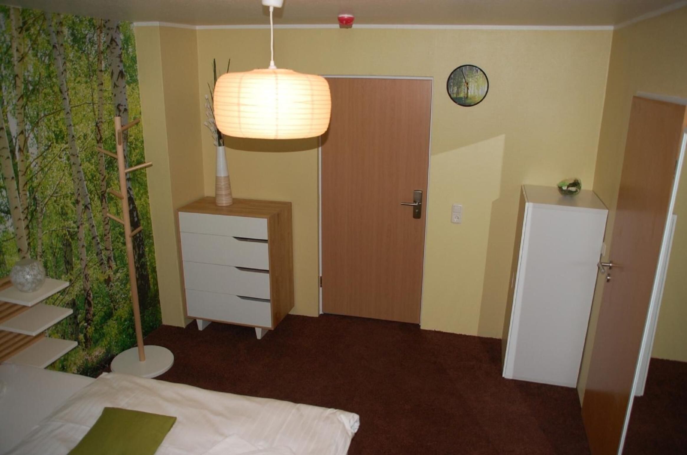 Un dormitorio con cama, cómoda, armario, mesita de noche, lámpara y un reloj de pared.