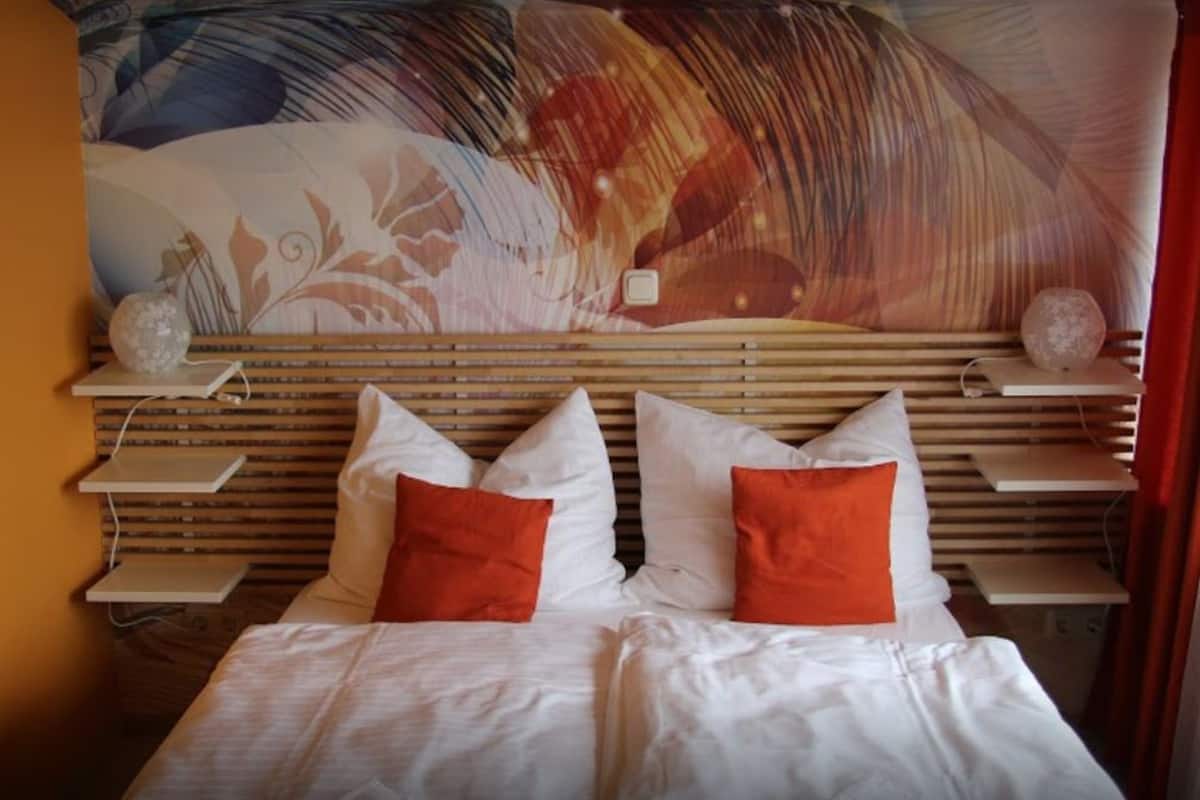 Ett hotellrum med en säng, vita sängkläder, två orangefärgade kuddar och en stor, abstrakt väggmålning.