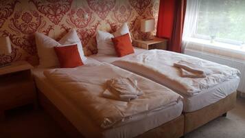 A double bed with white linens and orange pillows, a bedside table with a lamp, and a window with a view of greenery.
