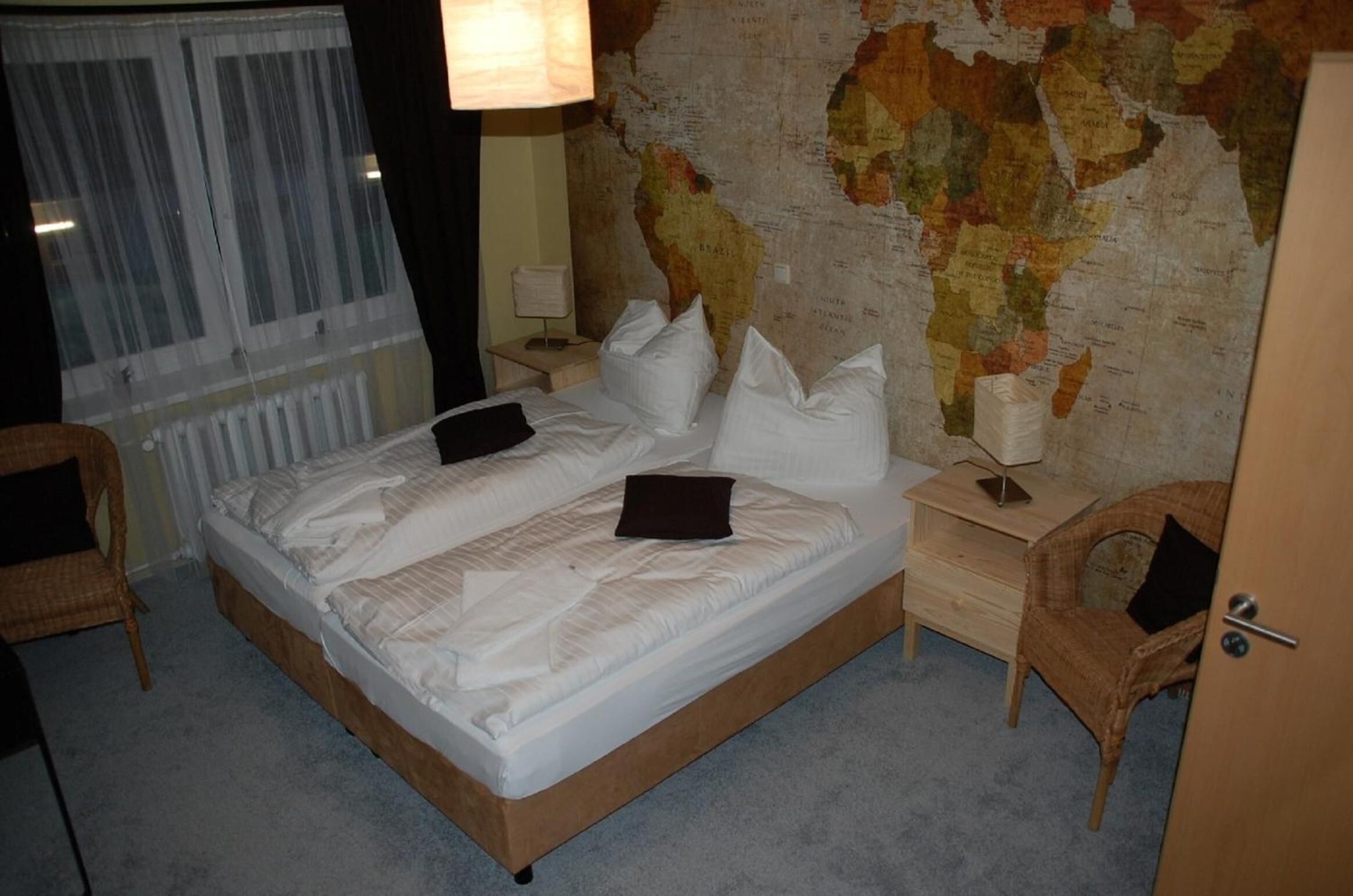 Un lit bien fait, avec des draps blancs et un plaid marron, une table de chevet en bois avec une lampe, une chaise en osier et une grande carte du monde accrochée au mur.