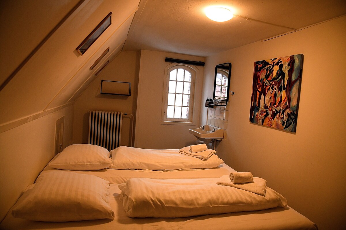 A bedroom with a bed, a radiator, a window, a sink, and a painting.