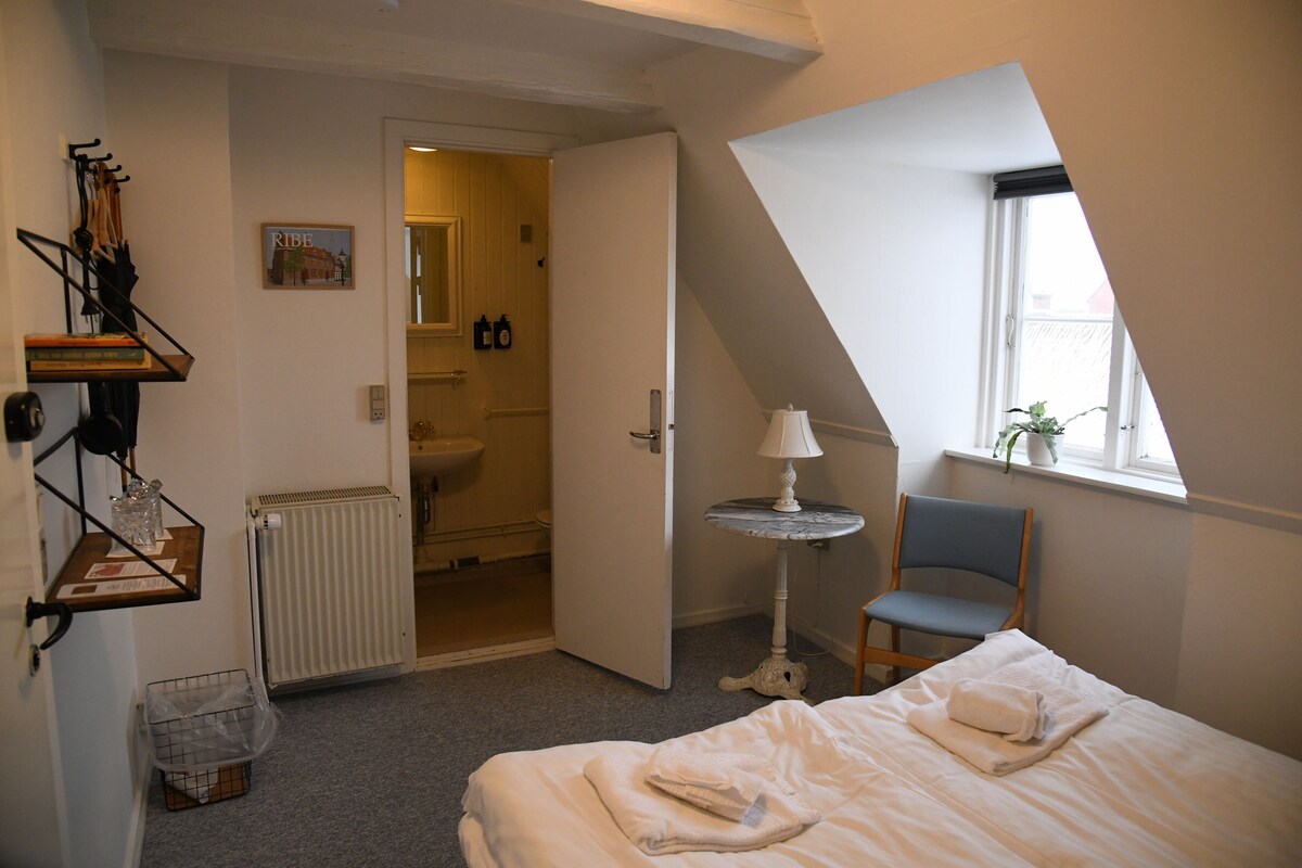 A bedroom with a bed, a chair, a lamp, a radiator, a window, and a door leading to a bathroom.