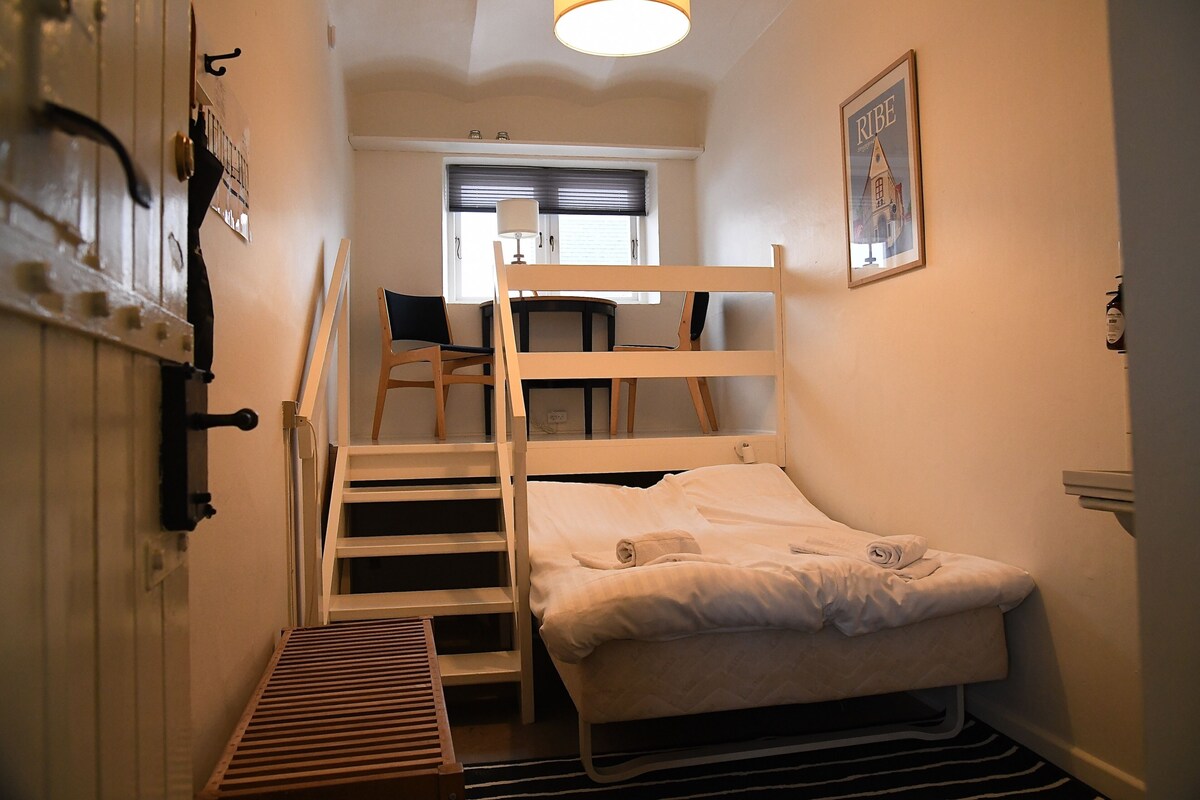 A small, compact room with a bunk bed, a desk, and a chair. There is a window with blinds, a radiator, and a framed picture on the wall.