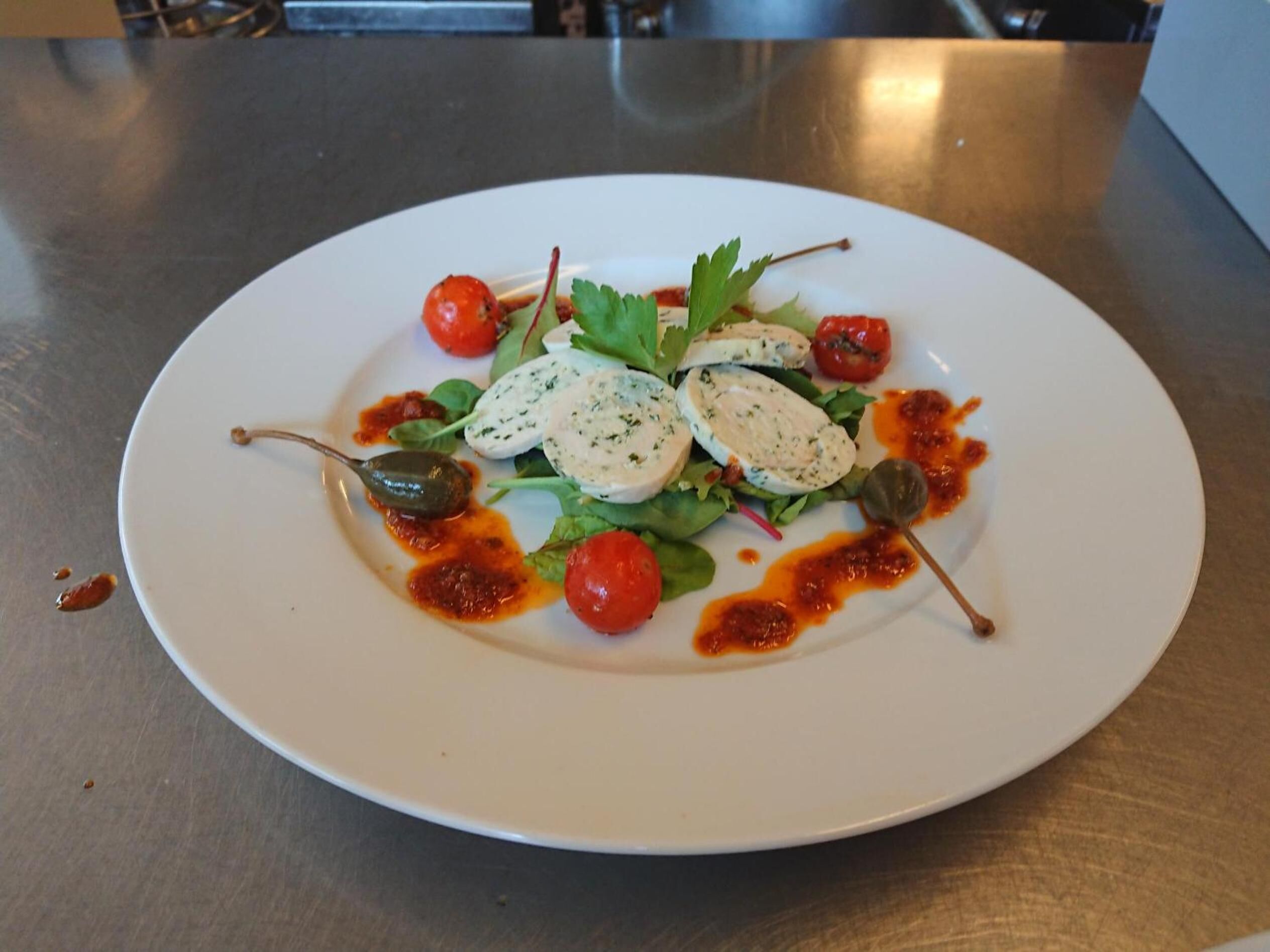 Un plato blanco con una ensalada, que incluye hojas verdes, tomates cherry y queso blanco en rodajas, sobre una mesa de acero inoxidable.